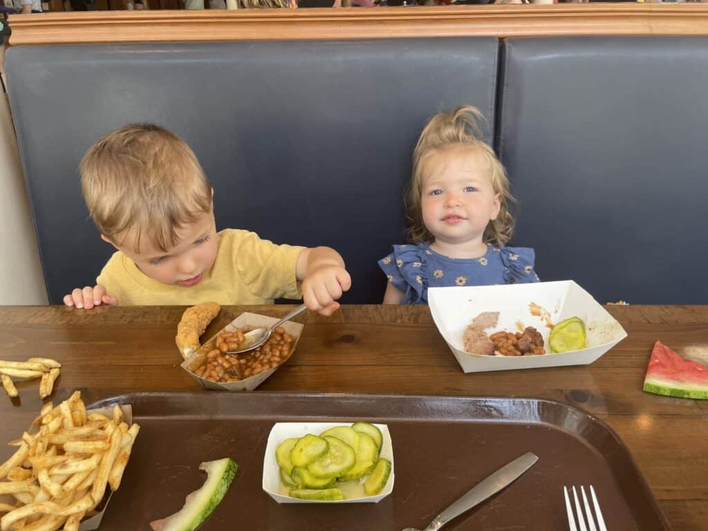 Disney World alone with toddlers: Young siblings enjoying a casual lunch at a restaurant booth, with baked beans, french fries, pickles and watermelon on the table. Boy in yellow shirt and girl in blue floral dress.