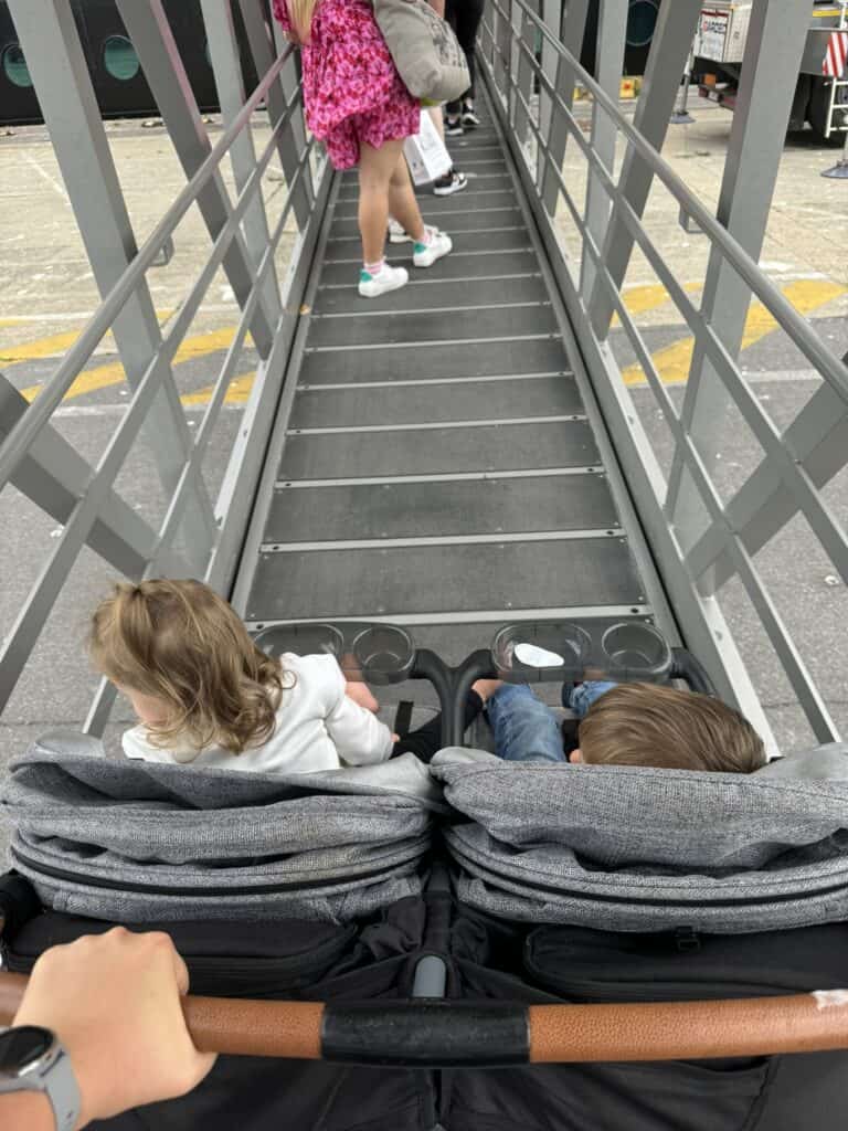 Two children are seated in a double stroller on a gangway leading to a cruise ship. A person in a pink dress is walking ahead, and the stroller is being pushed by someone with a watch.