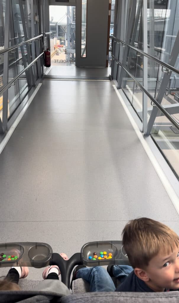 Two children are seated in a double stroller on a cruise ship gangway. The stroller trays have colorful candies, and the gangway is enclosed with glass walls.