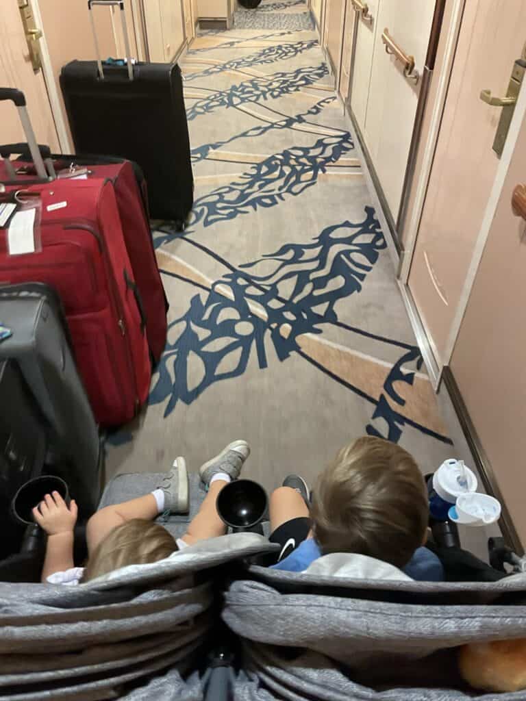 Hallway view aboard the Royal Caribbean Freedom of the Seas cruise ship, showing luggage against the wall and a double stroller passing easily beside it. The cruise ship carpet features an elegant blue and beige pattern, with cream-colored walls and brass handrails typical of cruise ship corridors.