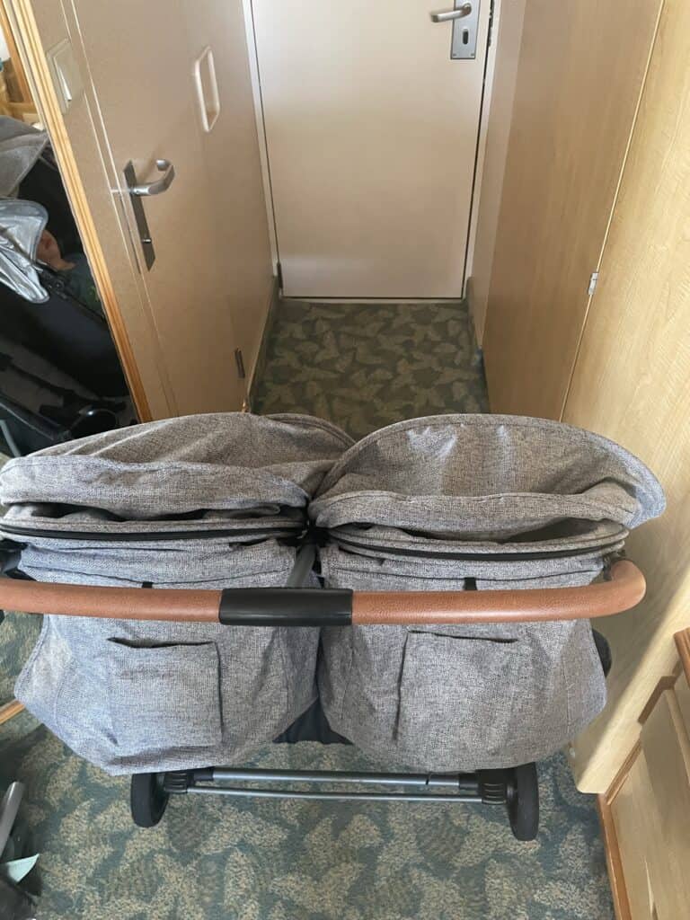 Close-up view of a gray double stroller on Royal Caribbean in a cstateroom entryway, just unable to fit. It's showing the detailed construction of the stroller seats with their gray fabric and tan leather handlebar. The ship's cabin doors and carpeting are visible in the background.