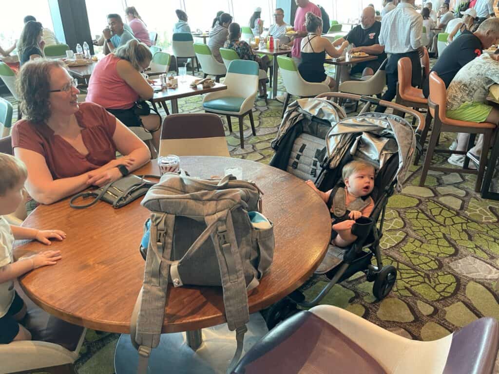The Windjammer buffet restaurant on a Royal Caribbean cruise ship, showing a round wooden table with a gray backpack and a double stroller pulled up to the table. The space features the cruise line's typical modern decor with mint green and white chairs, and their signature geometric-patterned carpet in green tones. The buffet area is busy with other cruise passengers dining.