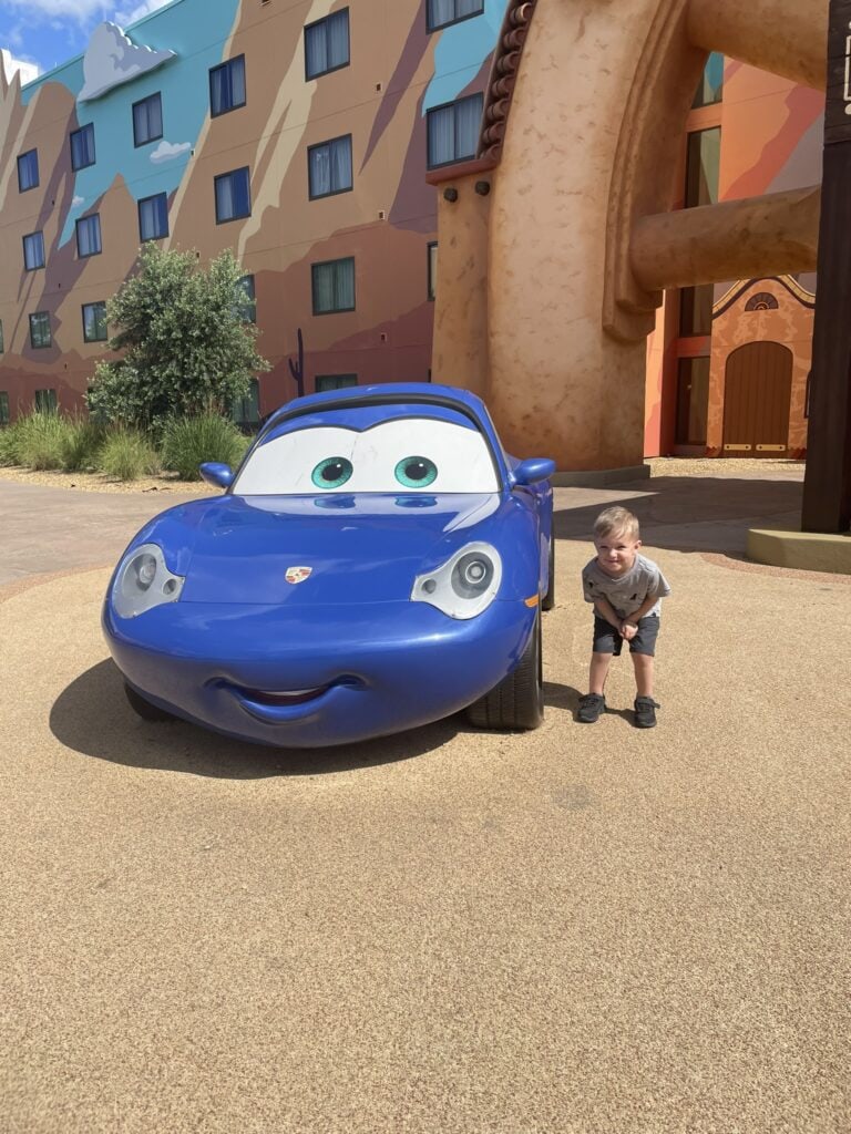 A small child squats down next to a blue car from the Disney movie Cars. There is a hotel in the background which is also themed to the movie.