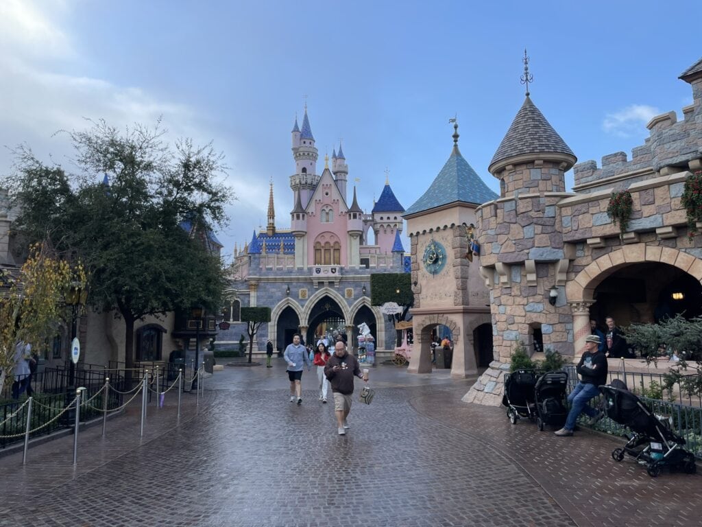 A view of Sleeping Beauty Castle at Disneyland Park on a damp, overcast day, with a few strollers parked along the right side, indicating Disneyland Park stroller parking. Guests walk along the cobblestone path leading to the pink and blue castle surrounded by whimsical towers and fairytale architecture.