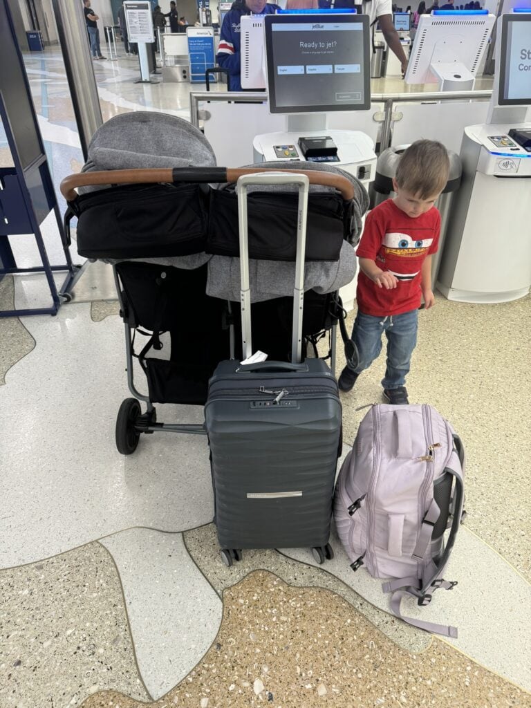 A gray double stroller, a rolling suitcase, and a backpack stand at an airport check-in kiosk. A young boy wearing a red Cars T-shirt stands beside them, looking down.