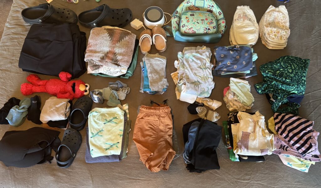 A complete flat lay of a suitcase's contents for a trip with toddlers, including shoes, diapers, clothes, toys, a sound machine, and a Disney Parks mini backpack—perfect for a visual packing list for traveling with toddlers.