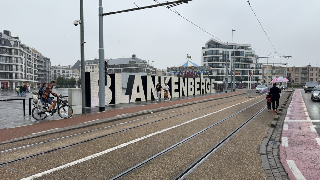 A large “BLANKENBERGE” sign stretches along the promenade near tram tracks, with a carousel behind and people walking and biking nearby. It’s a popular seaside stop during Belgian coastal visits.