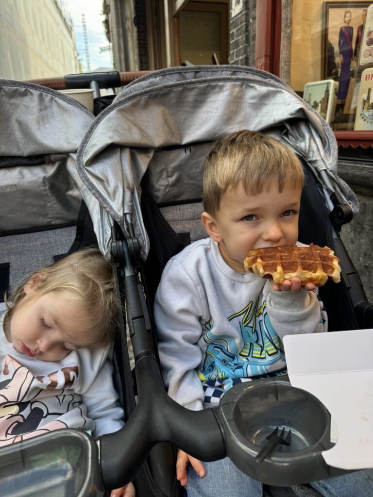 A young boy eats a Belgian waffle while sitting in a double stroller next to his sleeping sister, outside a shop in Bruges. The moment captures the charm of travel with toddlers (and snacks).