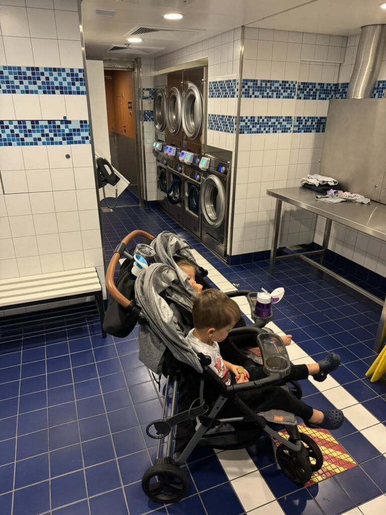 A double stroller with two toddlers sits near the front of the self-service laundry room onboard the ship, with washing machines, dryers, and folding tables in the background—useful for longer family cruises.