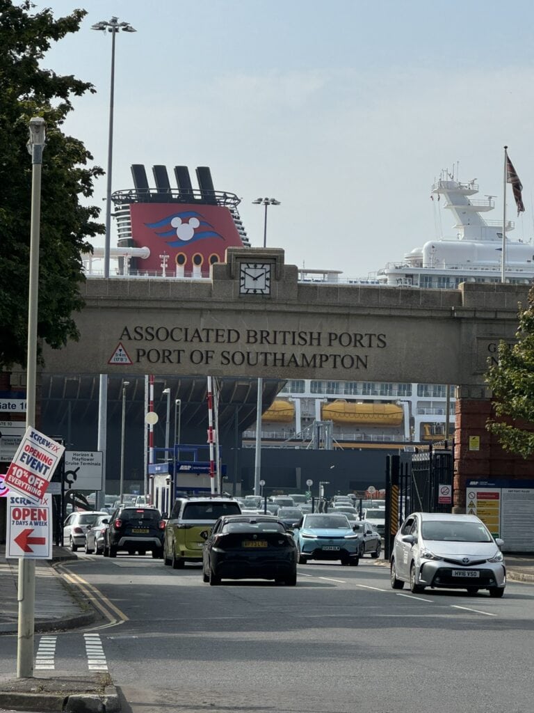 A view of the entrance to the Port of Southampton with a Disney Cruise Line ship docked in the background. The terminal area is bustling with cars, framed by the iconic Associated British Ports sign.