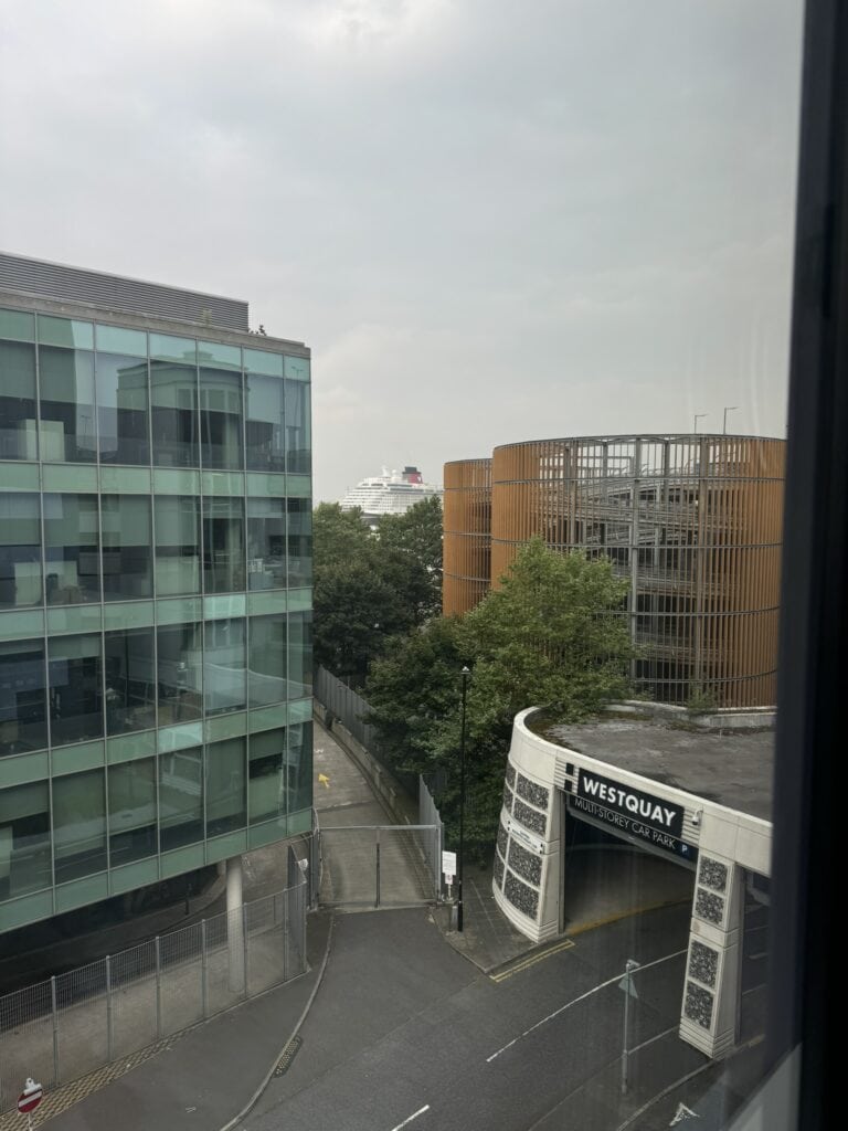 Looking out from a hotel window, the image shows the Westquay parking garage and nearby office buildings, with the red funnel of a cruise ship visible in the distance. A subtle peek at the cruise-ready location near central Southampton.