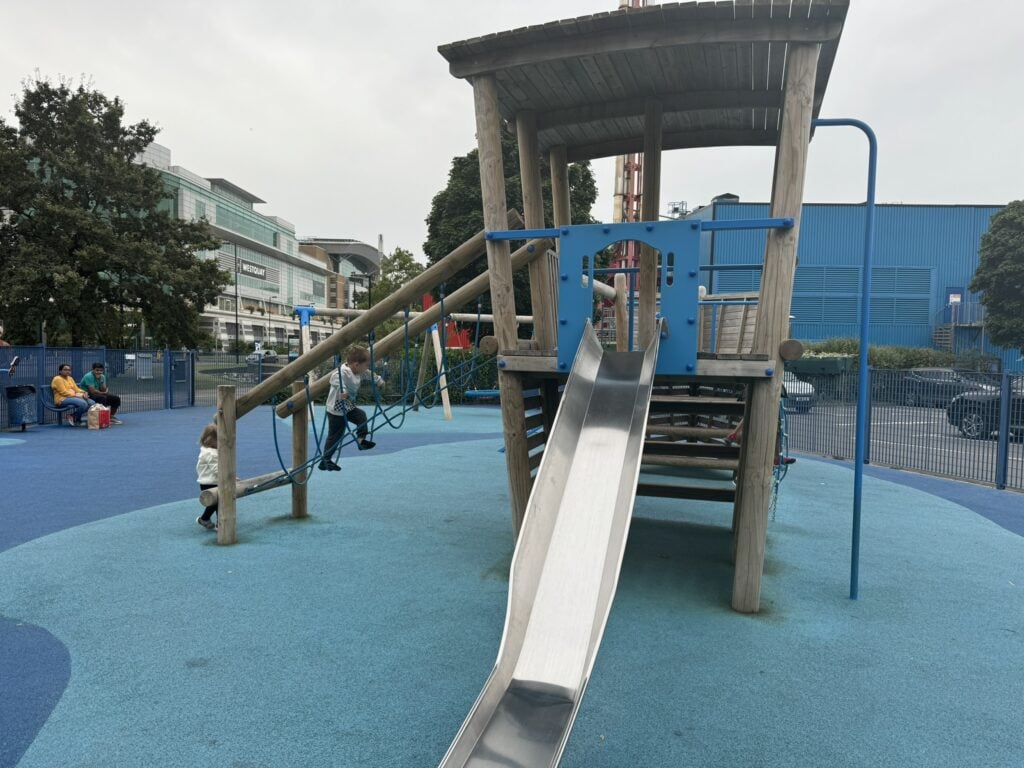 Two young children play on a modern playground with a rope climb and slide, located near the Westquay shopping center in Southampton. A great example of how easy it is to entertain little ones while staying at Moxy Southampton with kids.