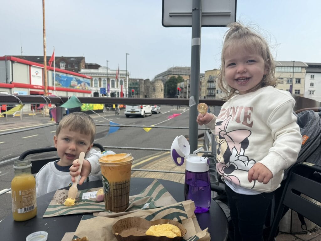Two kids enjoy breakfast at an outdoor Starbucks table near the Southampton waterfront, one with a cake pop and the other eating with a wooden fork. Juice bottles and coffee cups are on the table, with city buildings and cruise banners in the background.