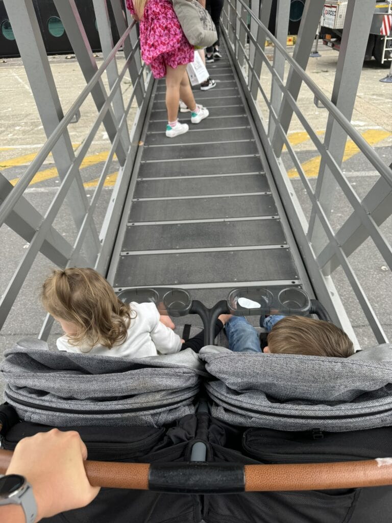 A parent pushes a double stroller with two toddlers up the gangway to board a Disney Cruise Line ship at the Zeebrugge cruise port. Other passengers walk ahead, with the ship's hull visible in the background.