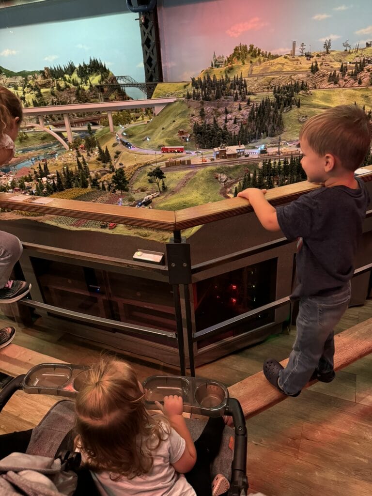 A toddler in a stroller and a young boy stand side by side, mesmerized by the detailed model landscape in front of them. This setup highlights the ease of visiting Miniatur Wunderland with a stroller, with stroller-friendly railings and tiered viewing platforms.