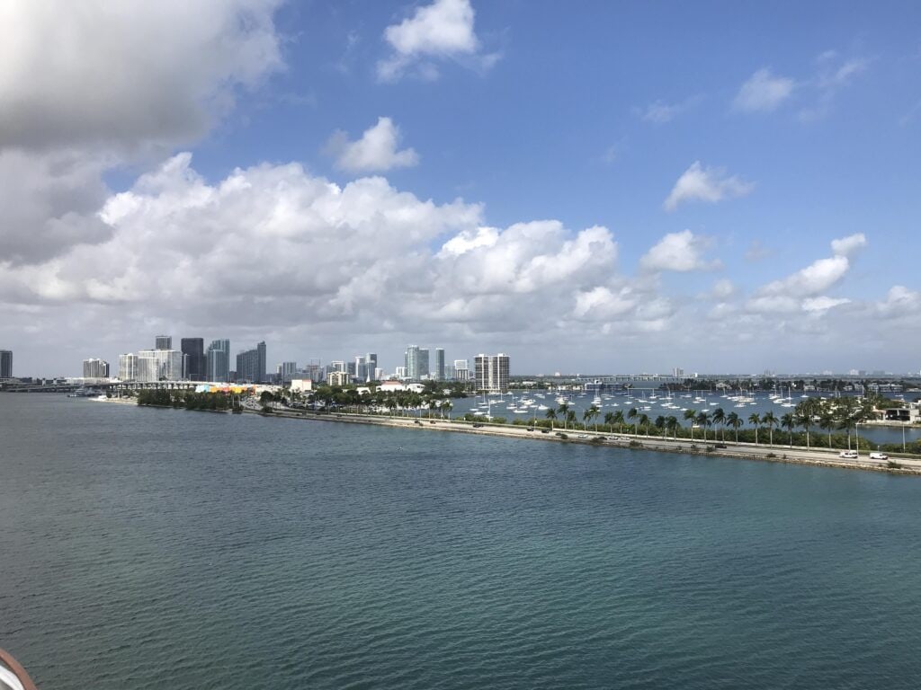 Sweeping view of Miami’s coastline with turquoise waters, palm-lined streets, and rows of anchored boats, with downtown skyscrapers visible in the distance under a partly cloudy sky.