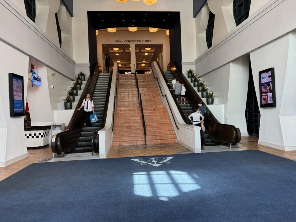 Guests ride up and down parallel escalators at the Dolphin Hotel’s main entrance, leading to the lobby and convention center, with restaurant signage visible below.
