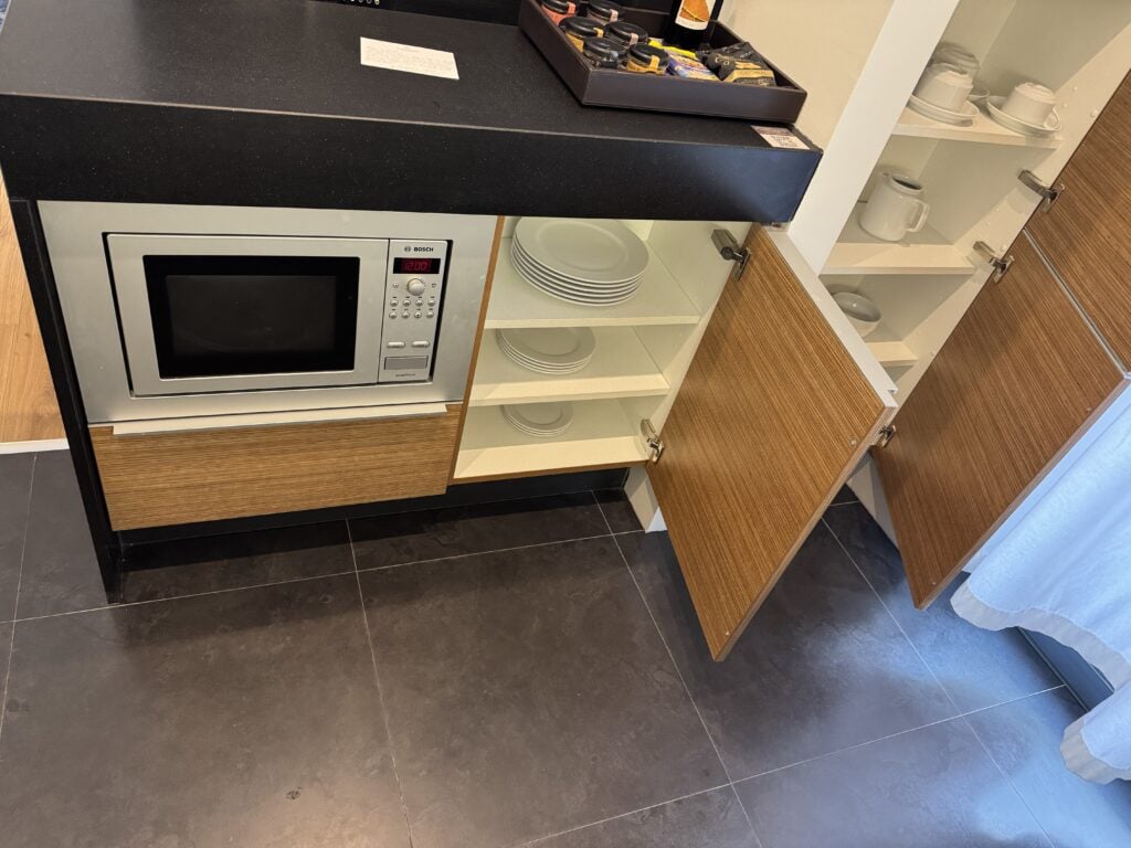 Open cabinet in a Sheraton Cascais Resort room showing glass cups, wine glasses, and a microwave all stored at toddler height—an inconvenient and potentially unsafe setup for families with young children.