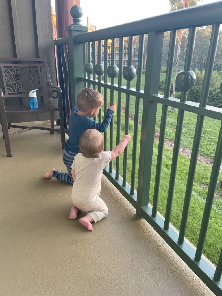Two young children in pajamas kneel on a balcony at Animal Kingdom Lodge, peeking through the railings at the savanna view below.