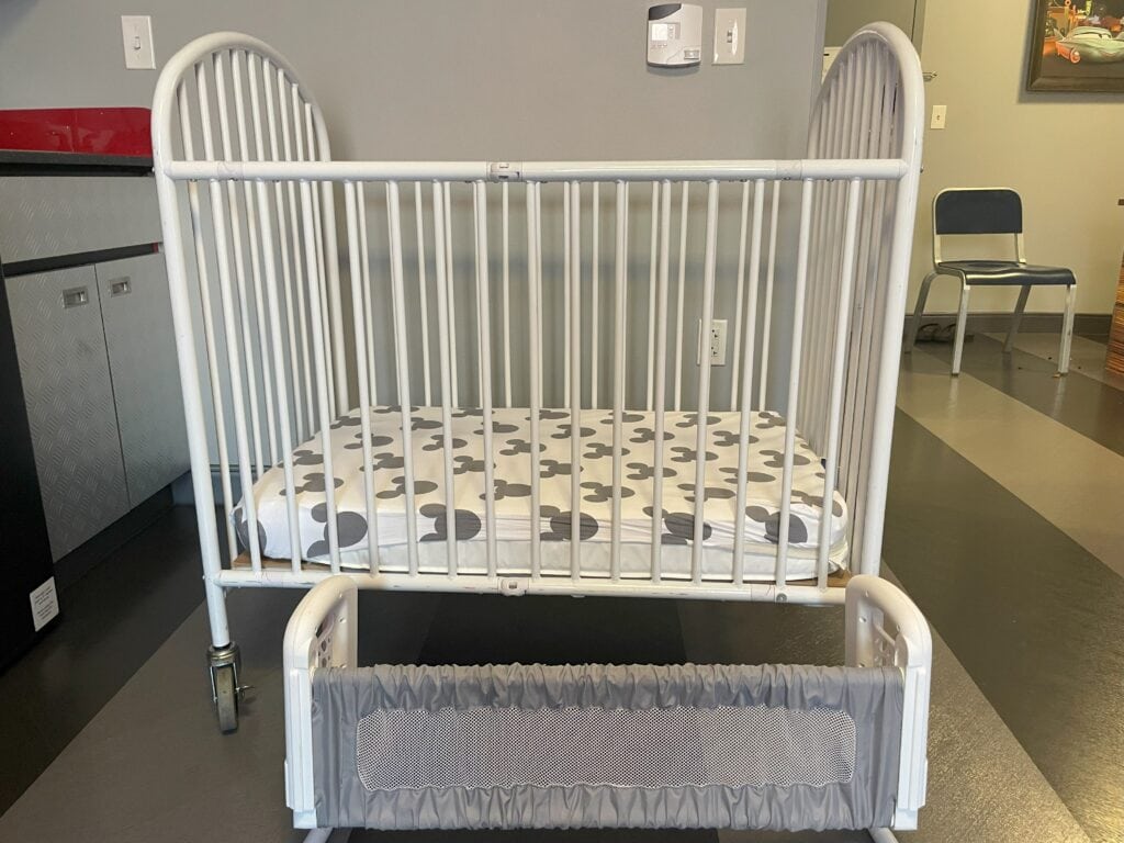 White metal crib with gray and white Mickey Mouse sheets, shown next to a gray bed rail provided by the Disney resort—perfect for keeping toddlers safe during sleep.