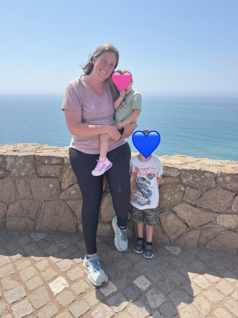 A smiling woman stands with two young children in front of a stone wall overlooking the ocean at Cabo da Roca, the westernmost point of Europe.