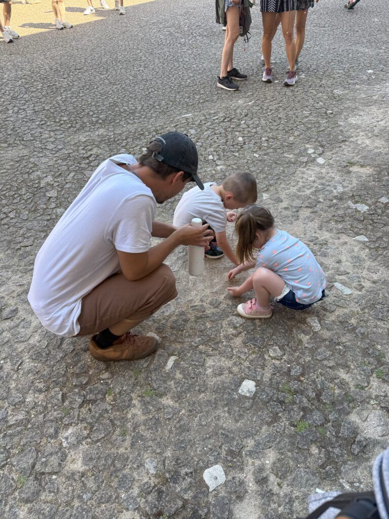 A man kneels next to two toddlers playing on cobblestone, taking a water break during a guided tour of Pena Palace.