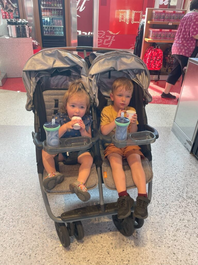 Two toddlers sit side-by-side in a Zoe Twin+ V1 stroller inside Epcot’s Club Cool, sipping Coca-Cola samples from small paper cups. The stroller trays hold their water bottles, and the bright red Coca-Cola display forms the backdrop.