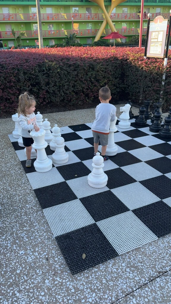 Two toddlers playing on a giant outdoor chessboard at Disney’s All Star Music Resort, surrounded by colorful buildings decorated with musical notes.