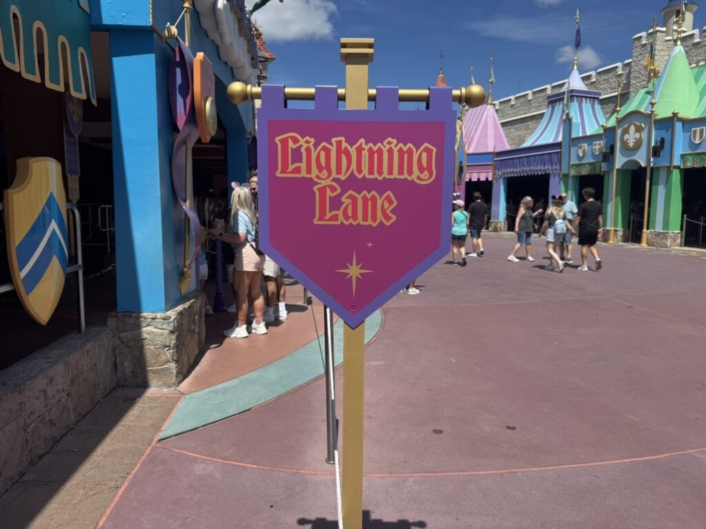 Purple Lightning Lane sign in front of Peter Pan’s Flight in Fantasyland at Magic Kingdom, with colorful medieval-style façades in the background.