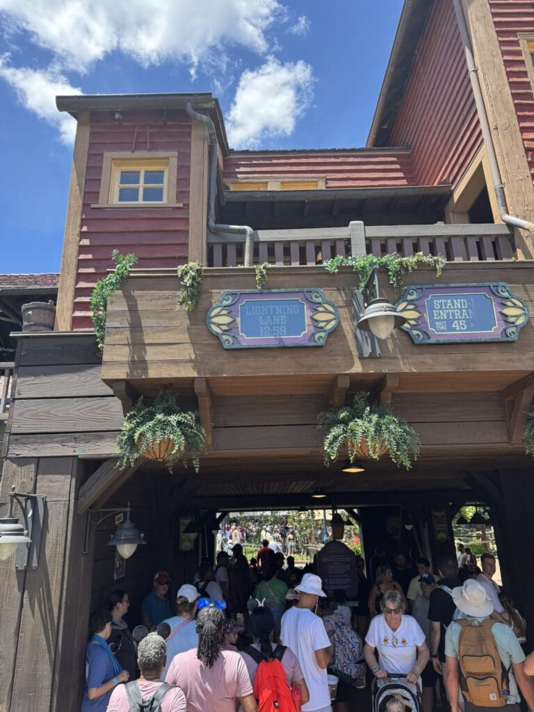 Crowds gathered under the entrance to Tiana’s Bayou Adventure at Magic Kingdom, with Lightning Lane and Stand-By wait time signs overhead.