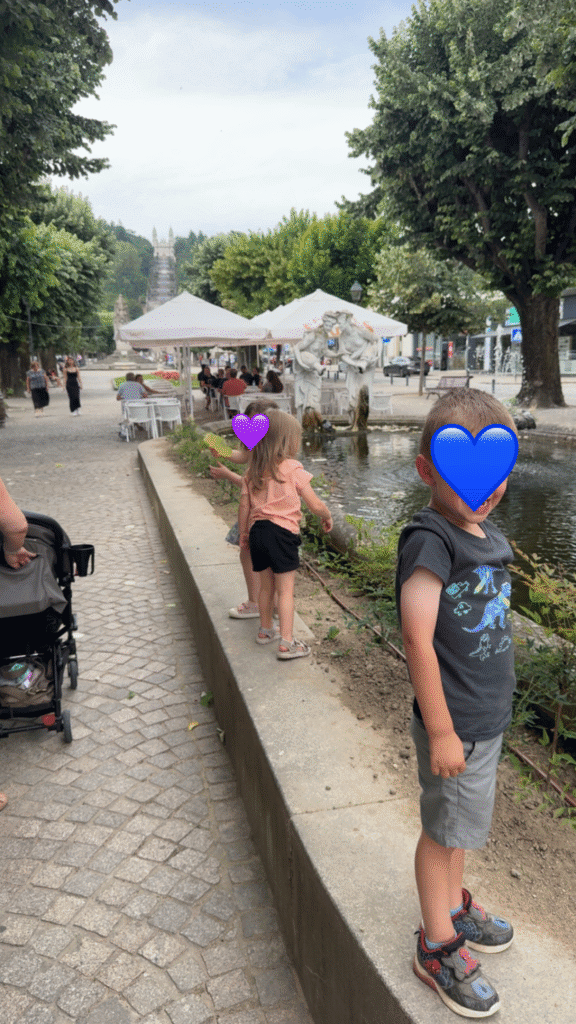Children play near a fountain and pond along a shaded pedestrian street in Lamego with families dining at outdoor cafés in the background. Perfect snapshot of exploring Lamego with kids.