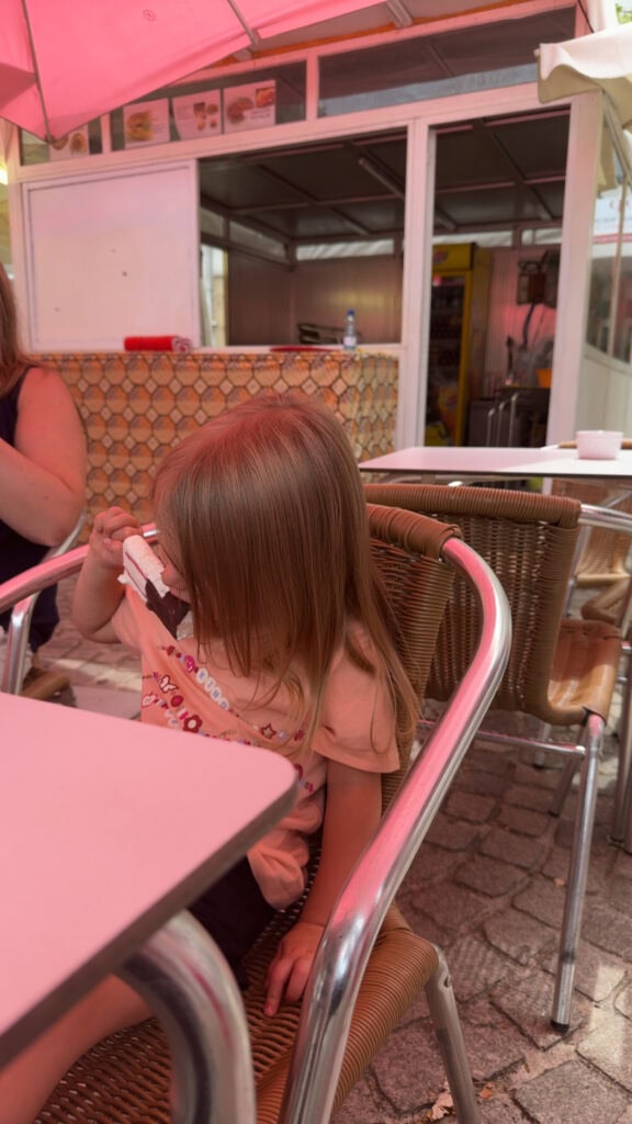 Young girl in a pink shirt sits at a metal café table under a red umbrella in Lamego, enjoying an ice cream treat.
