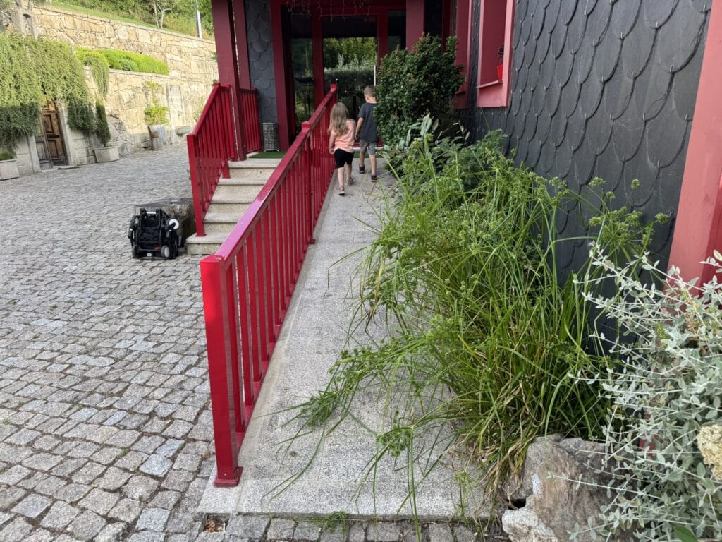 Two children walk up an overgrown accessibility ramp with bright red railings at Quinta Dos Padres Santos, with a folded stroller resting nearby.