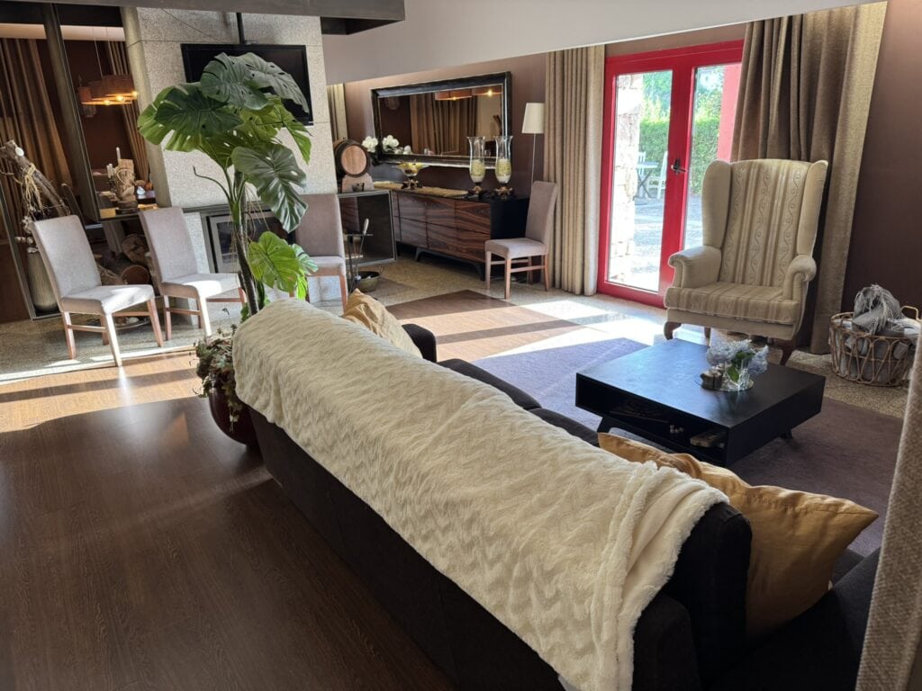 Elegant common room with a sofa draped in a cream throw, tall potted plant, armchairs, and warm natural light streaming through red-framed glass doors.