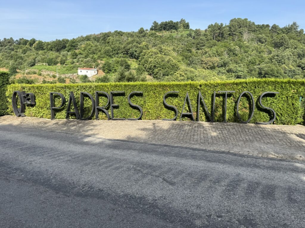 Large black metal letters spelling “Qta. Padres Santos” along a neatly trimmed hedge, with a hillside and white farmhouse in the background.