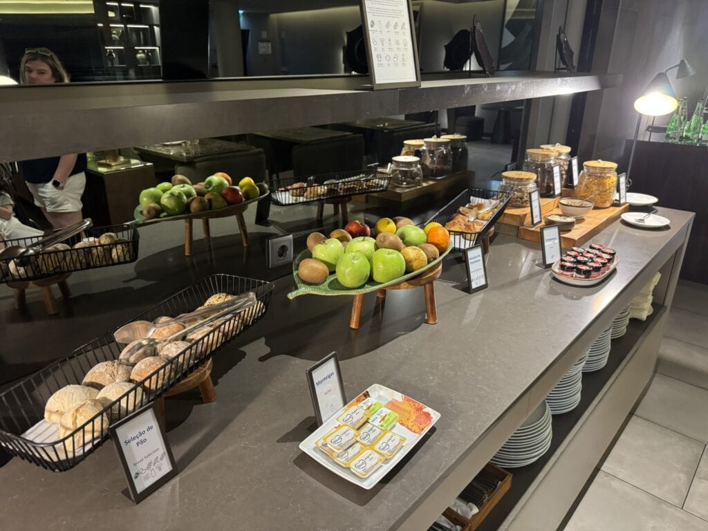 Close-up of the breakfast options in the executive lounge at Hilton Porto Gaia with fresh apples, bread rolls, butter, jams, cereal jars, and juice glasses arranged on a long countertop near mirrored walls.