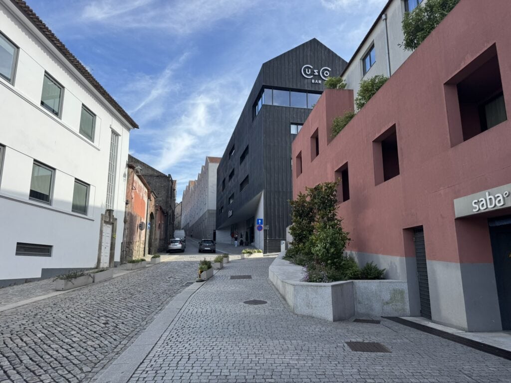 Sloped cobblestone street lined with white, red, and modern black buildings near Hilton Porto Gaia, with the Cusco Bar sign visible under a bright blue sky.