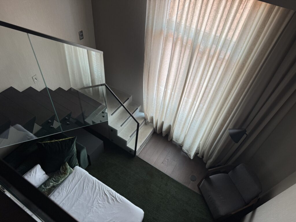 Interior view of the two-story Family Room suite at Hilton Porto Gaia with kids, showing the glass-paneled staircase, floor-to-ceiling curtains, dark green carpet, and lower-level sleeping and lounge area.