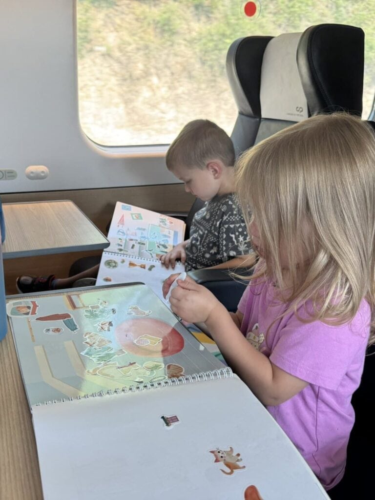 Two toddlers sit at a train table in a modern high-speed train, focused on sticker books. A pink-and-blue gradient water bottle marked with motivational time goals is on the table next to them.