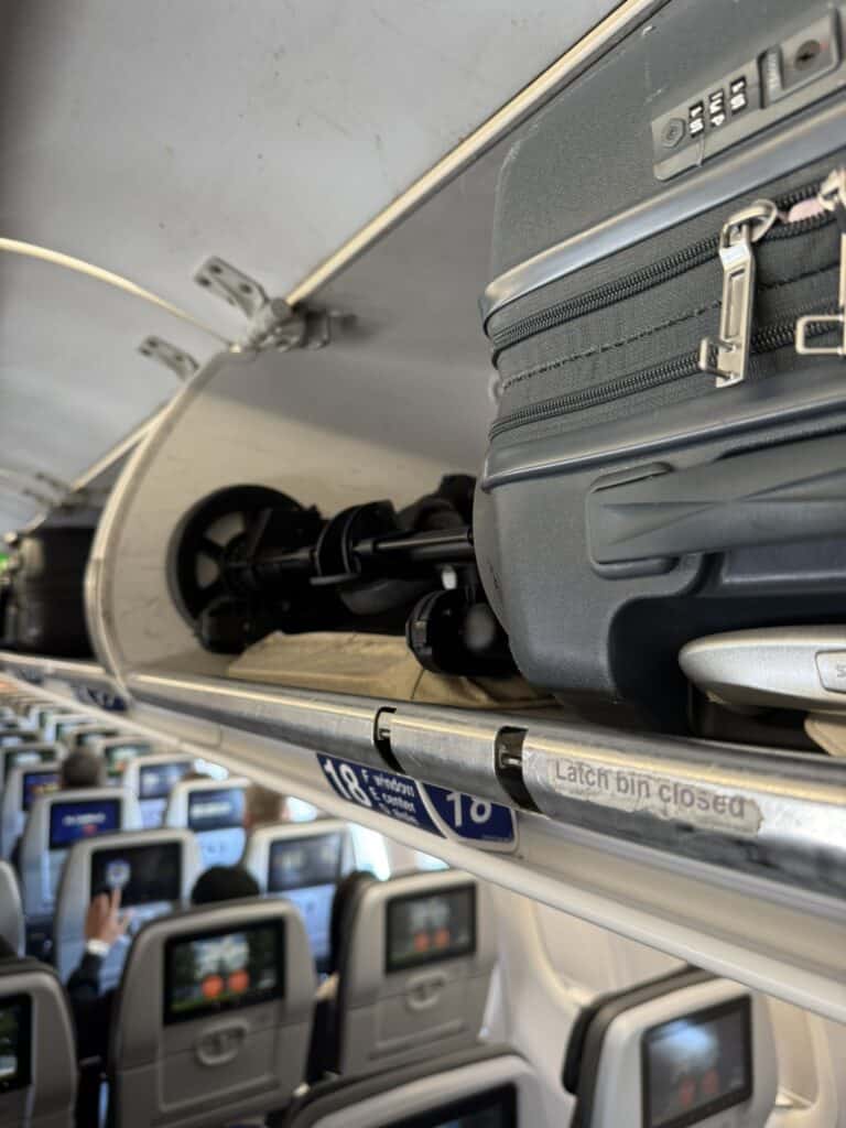 Folded Zoe Traveler stroller stored in an airplane overhead bin next to a carry-on suitcase during a JetBlue flight to Orlando.